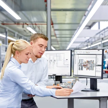 Ein Mann und eine Frau schauen auf einen Schaltplan. Auf dem Arbeitsplatz stehen zwei Monitore mit einem Schaltplan und einem Klemmenleistenmodell darauf.